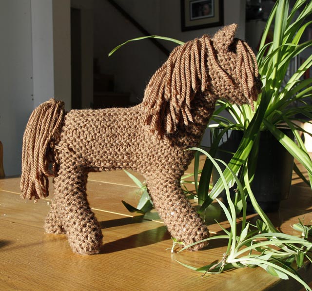 knit brown horse standing in the sunlight by a green spider plant